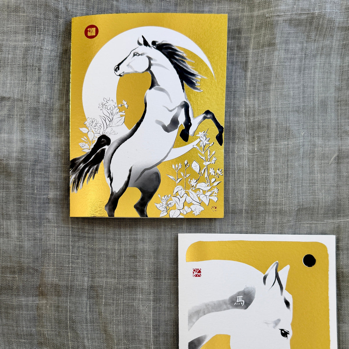 Two gold foil art cards featuring white horses on a textured gray fabric background. The top card shows a rearing horse against a crescent moon, and the bottom card shows a close-up of a white horse's head with a gold foil accent.