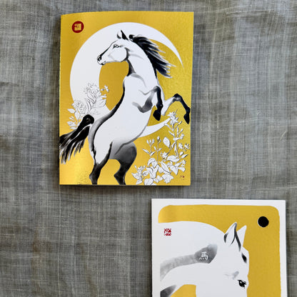 Two gold foil art cards featuring white horses on a textured gray fabric background. The top card shows a rearing horse against a crescent moon, and the bottom card shows a close-up of a white horse's head with a gold foil accent.