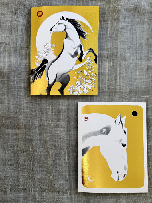 Two gold foil art cards featuring white horses on a textured gray fabric background. The top card shows a rearing horse against a crescent moon, and the bottom card shows a close-up of a white horse's head with a gold foil accent.